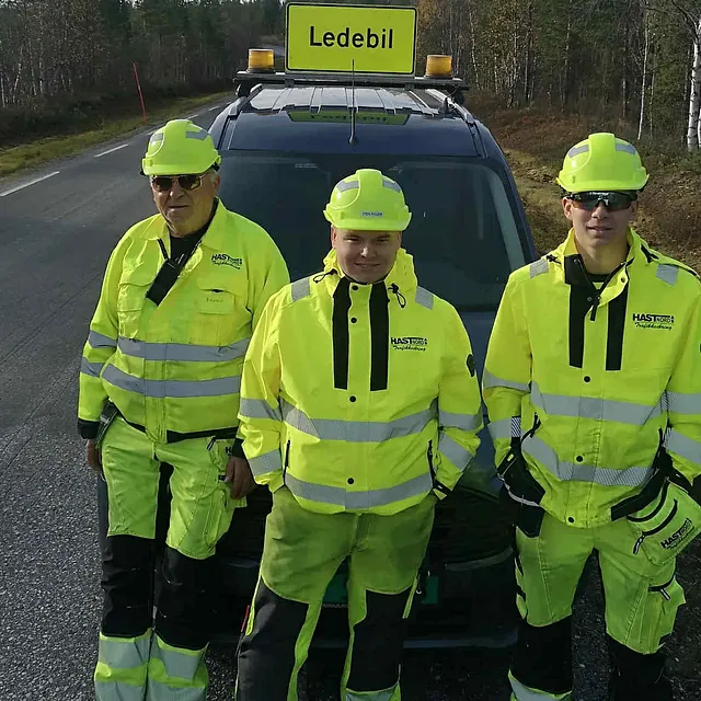 3 personer med gule refleksdresser, foran lederbil.